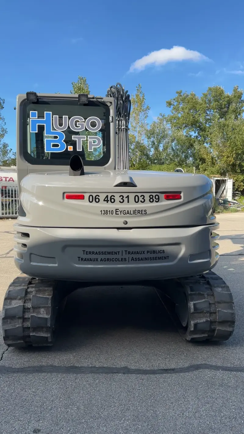 Arrière d'une pelle Hugo BTP floquée avec le logo, le numéro 06 46 31 03 89 et la mention Eygalières